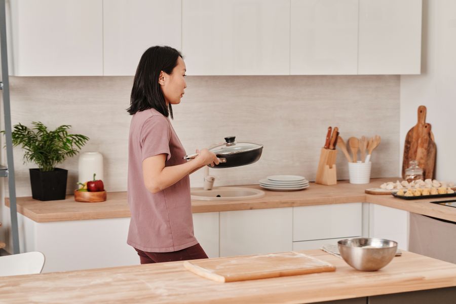Asian woman cooking in a modern kitchen holding a pan, surrounded by kitchen utensils.
