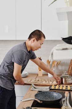 Asian man making homemade dumplings on a kitchen counter with a frying pan nearby.