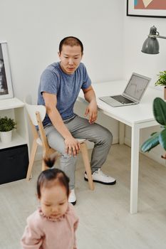 A father watching his daughter play near his workspace with a laptop, showcasing family bonding.