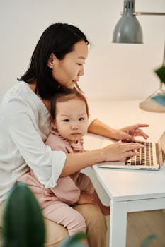 Asian mother multitasking with laptop and child at home.