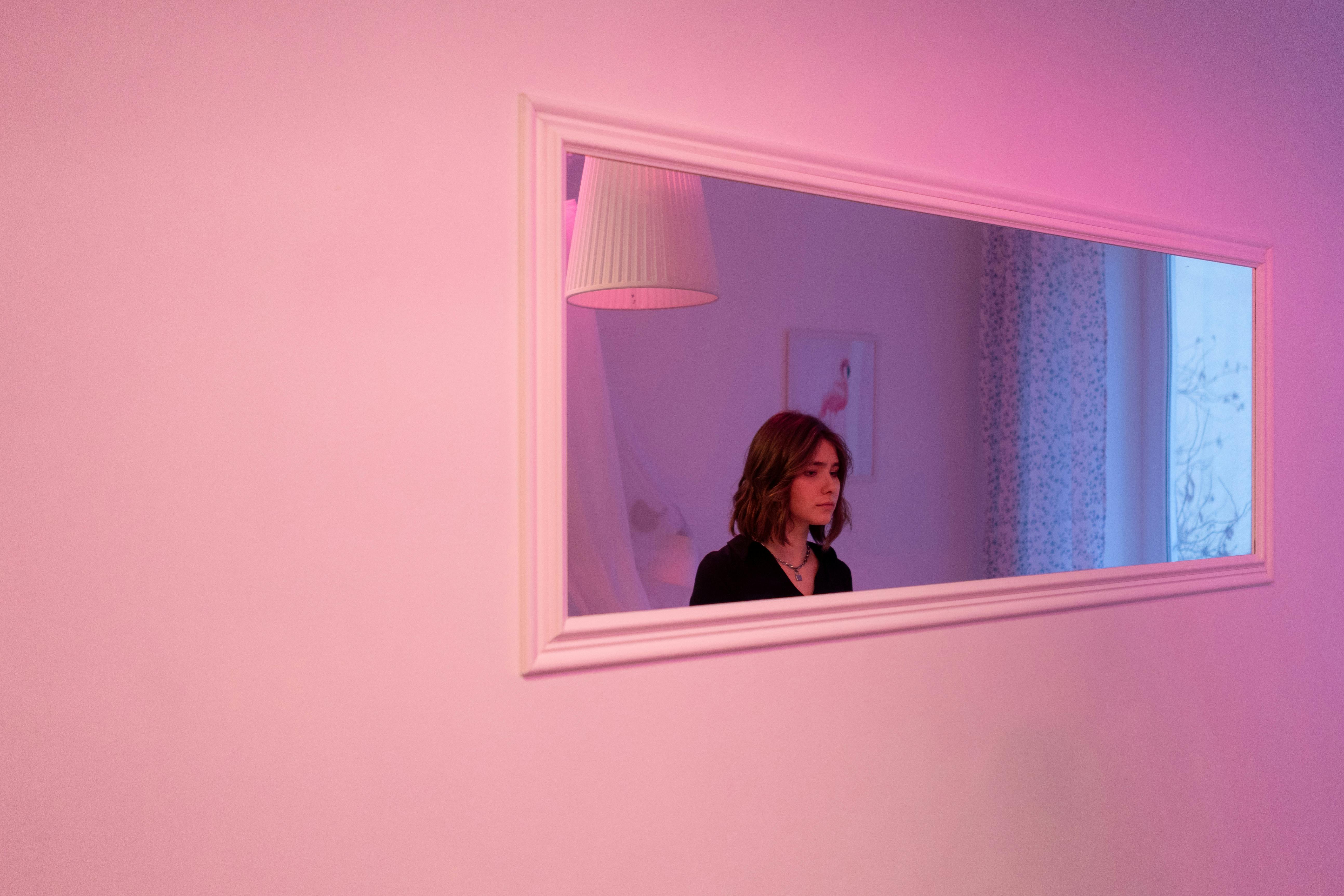 A woman with short hair reflects in a modern mirror under soft pink lighting.