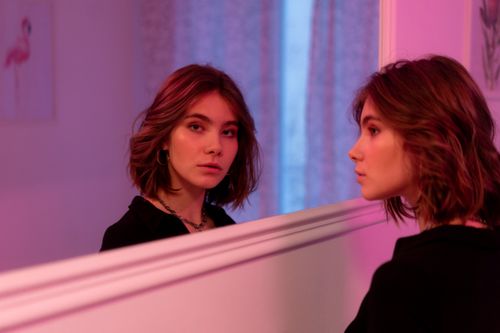 Free Young woman with short hair gazing at herself in a mirror, illuminated by soft pink lighting. Stock Photo