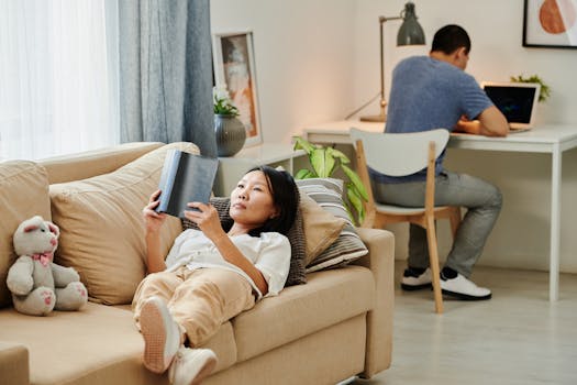 A woman reading on a sofa while a man works remotely at a desk in a cozy home setting.