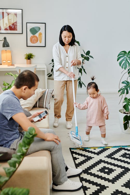 A Woman Using a Mop with Her Child while Her Husband is Sitting on a Couch