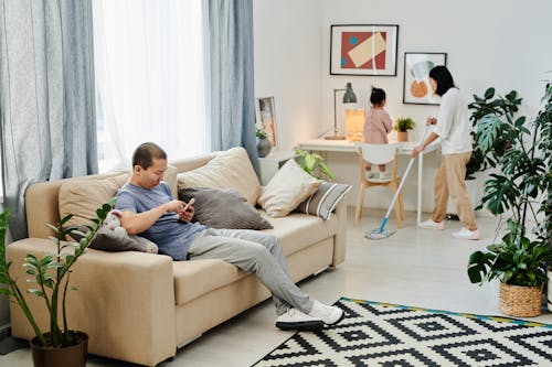 Wife Cleaning the Floor while Husband is Sitting on a Sofa with a Smart Phone