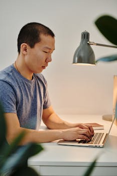 Man working remotely on a laptop at a desk with a lamp, showcasing a modern work-from-home setup.