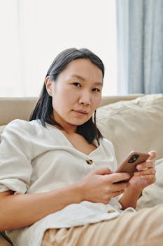 Asian woman comfortably using smartphone on a cozy sofa during the day.
