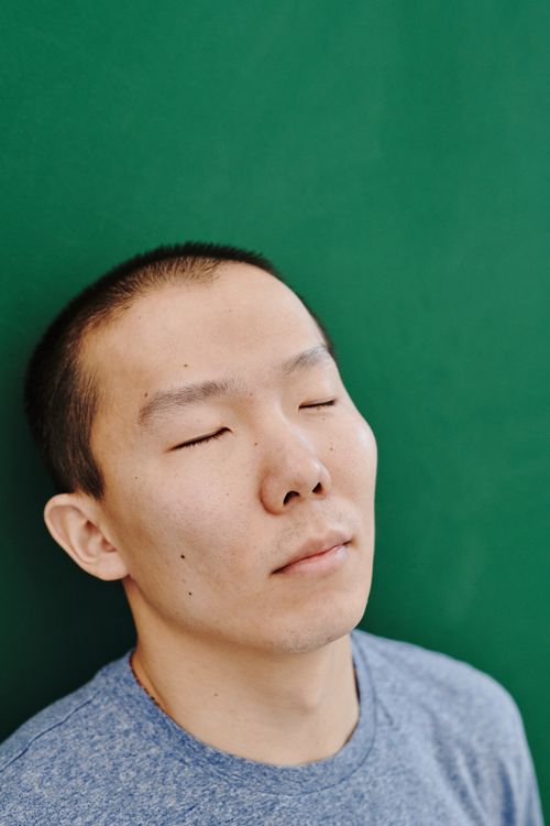 Free Close-up portrait of an Eastern Asian man with eyes closed, relaxing against a green wall. Stock Photo