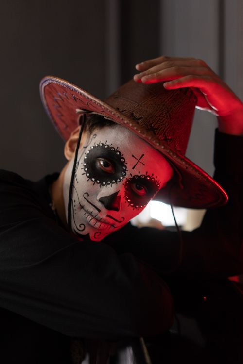 Free A dramatic portrait of a person wearing intricate Halloween face paint and a cowboy hat, lit with moody red lighting. Stock Photo
