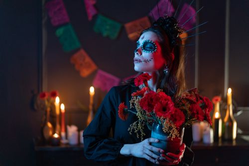 Free Woman with Day of the Dead makeup holding flowers, surrounded by candles and papel picado decorations. Stock Photo