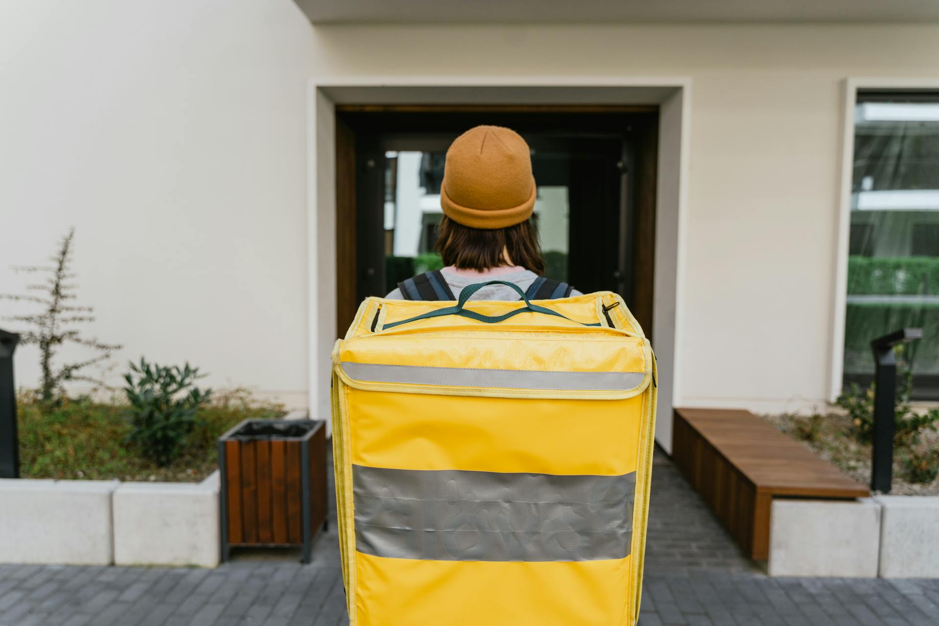 Rear view of a food delivery courier with a thermal backpack approaching a modern building entrance.