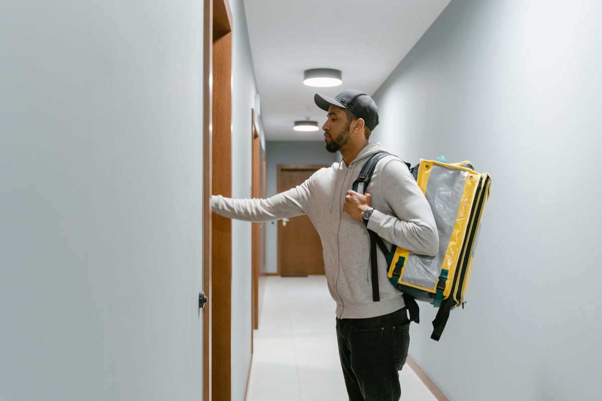 Courier wearing a backpack delivering food to an apartment door indoors.