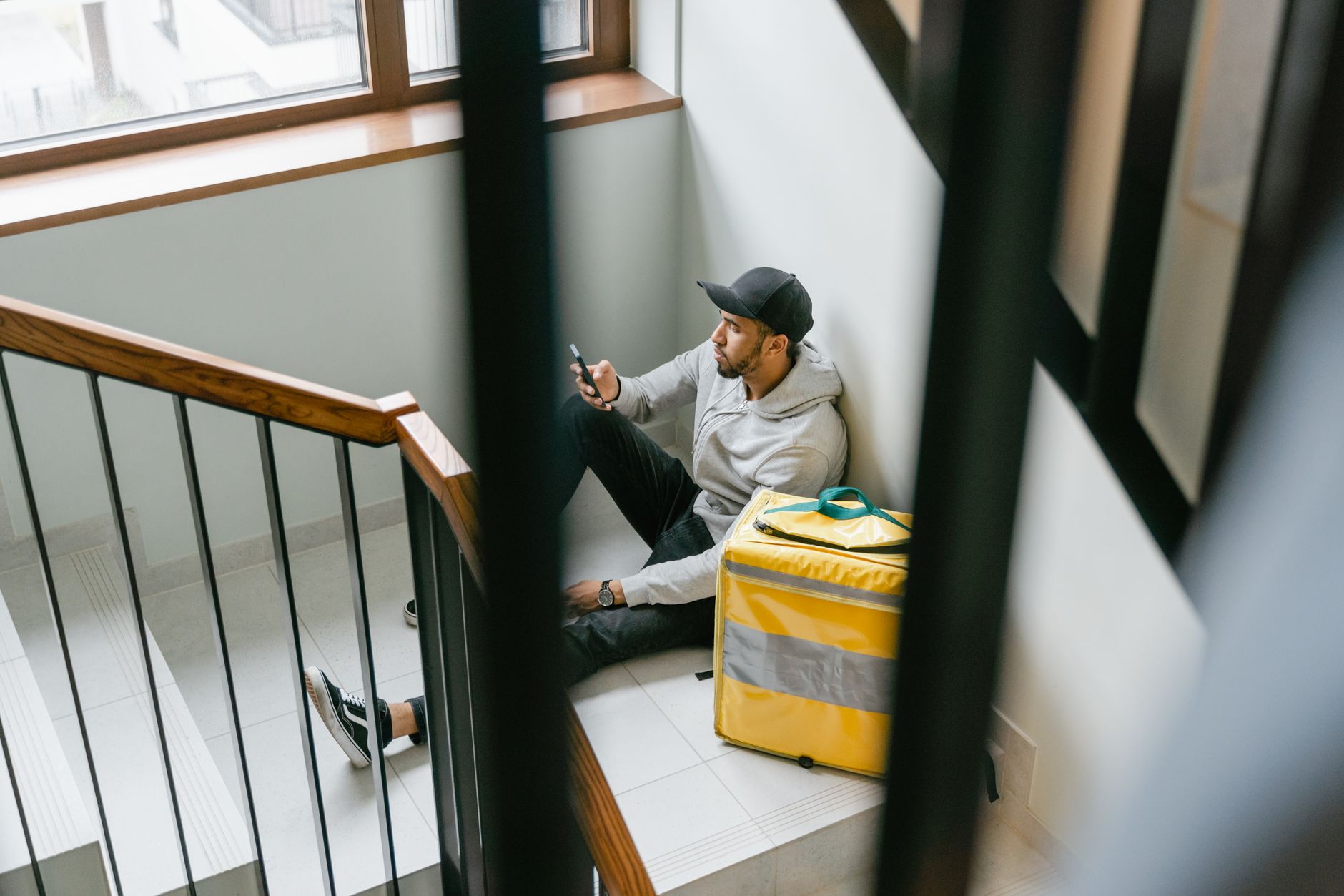 A food delivery man in casual attire sits on a staircase with a large yellow thermal bag.