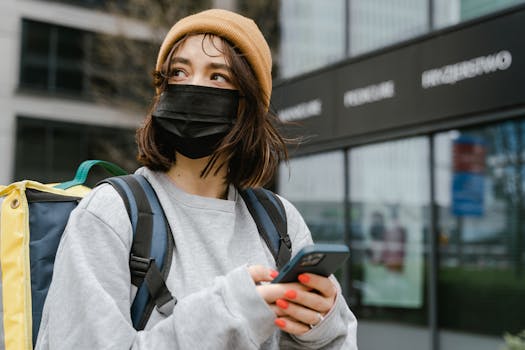 A female courier wearing a mask checks her smartphone in an urban setting, ready for delivery.