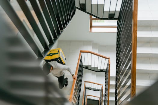 A delivery person carrying a yellow thermal bag ascends a modern staircase indoors.