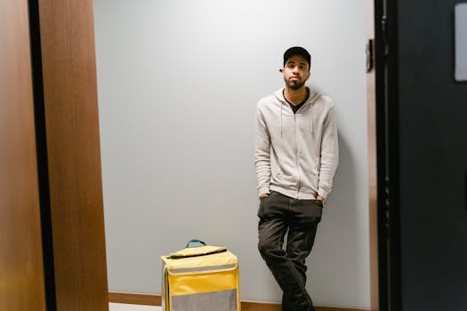 Young male delivery person waiting indoors with a yellow thermal bag, ready for food delivery.