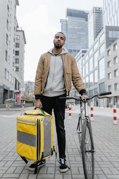 A bicycle courier holding a vibrant yellow thermal bag in an urban setting, ready for food delivery.