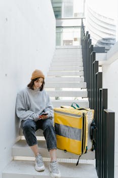 Woman courier with a yellow delivery bag on urban stairs, checking phone.