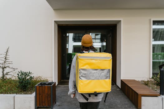 A food delivery person with a yellow thermal backpack entering a modern building.