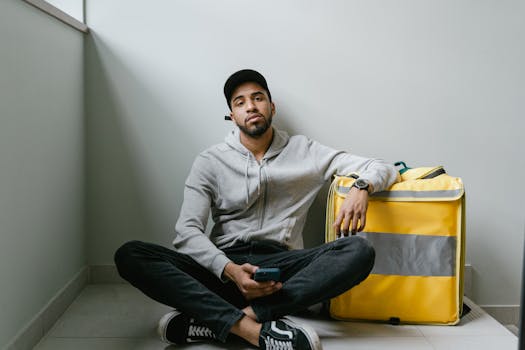 A food delivery courier sits on the floor, waiting with a yellow thermal bag inside a building.