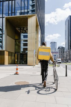 A city courier with a bicycle and delivery bag walking past modern skyscrapers.