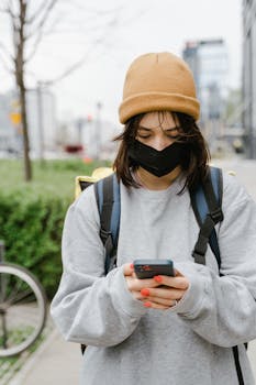 A delivery person wearing a mask and beanie checks their smartphone in an urban environment.