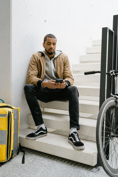 Free Young male food courier resting on stairs with bicycle and delivery bag nearby, checking phone. Stock Photo