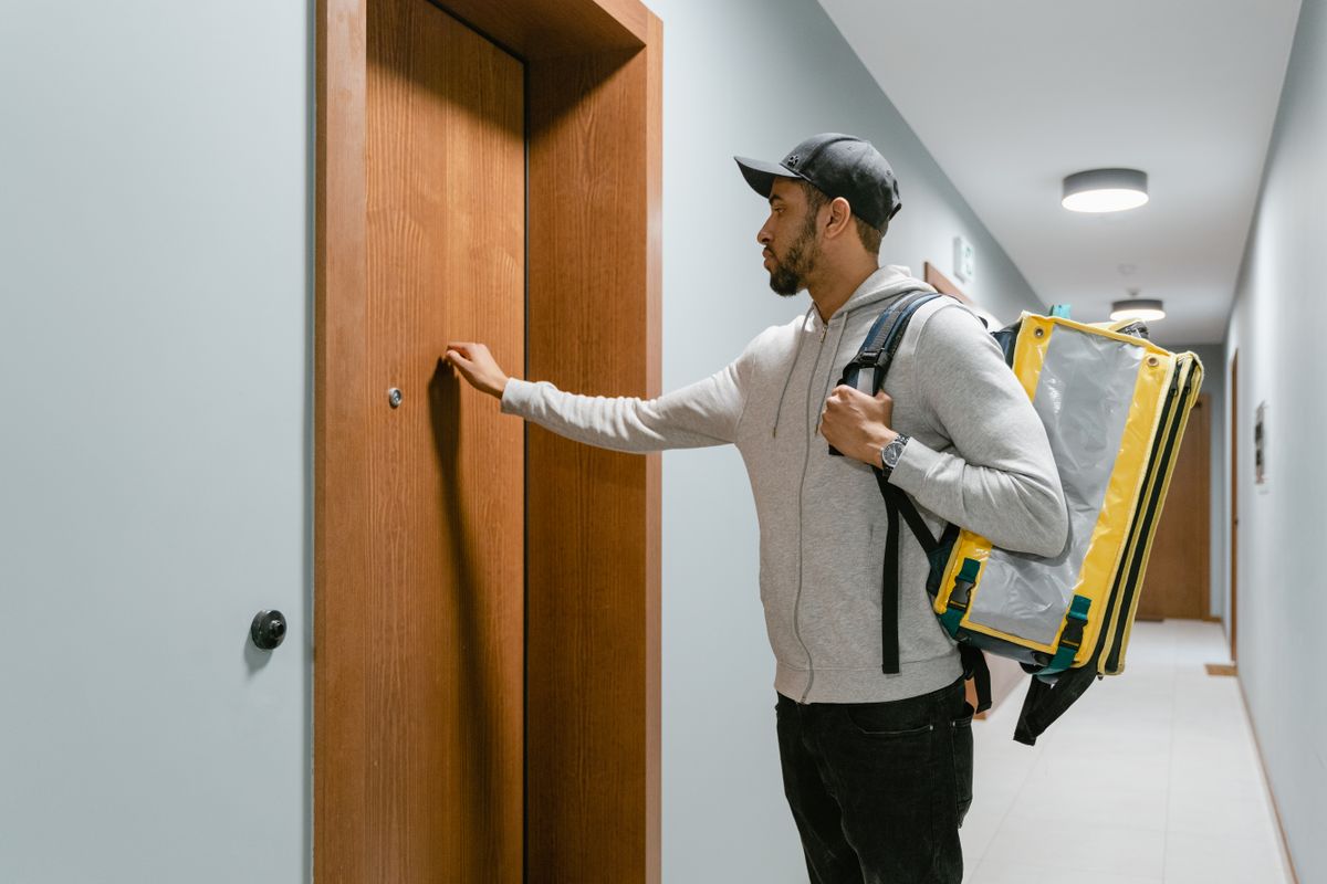 A food delivery man knocks on an apartment door, carrying a thermal bag.
