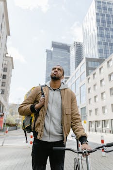 A cyclist with a backpack stands with his bicycle in a modern urban setting, looking upwards.