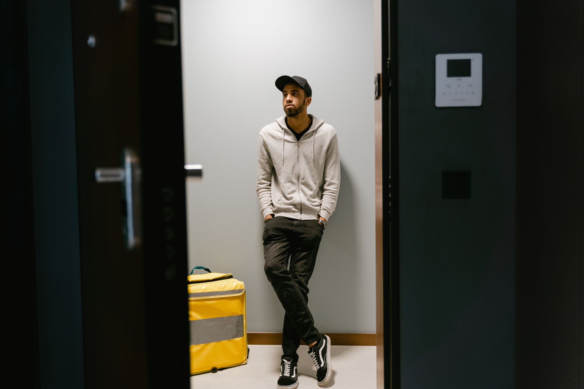 A man in casual attire stands waiting in a hallway with a delivery bag.