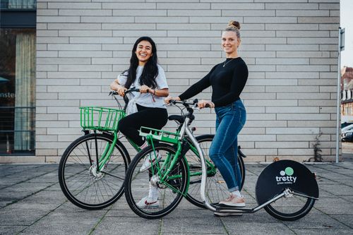 Free Two women smiling while riding bicycles and a kick scooter outdoors on a sunny day. Stock Photo