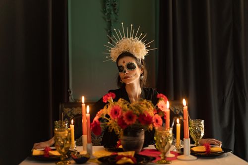 Free A woman adorned in Day of the Dead makeup and attire sits at a decorated table with candles and flowers, creating a festive atmosphere. Stock Photo