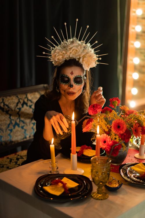 Free Woman with sugar skull makeup celebrates Day of the Dead with candles and flowers indoors. Stock Photo