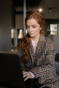 Caucasian woman with a checkered coat intently working on a laptop in an office setting. Professional and modern.