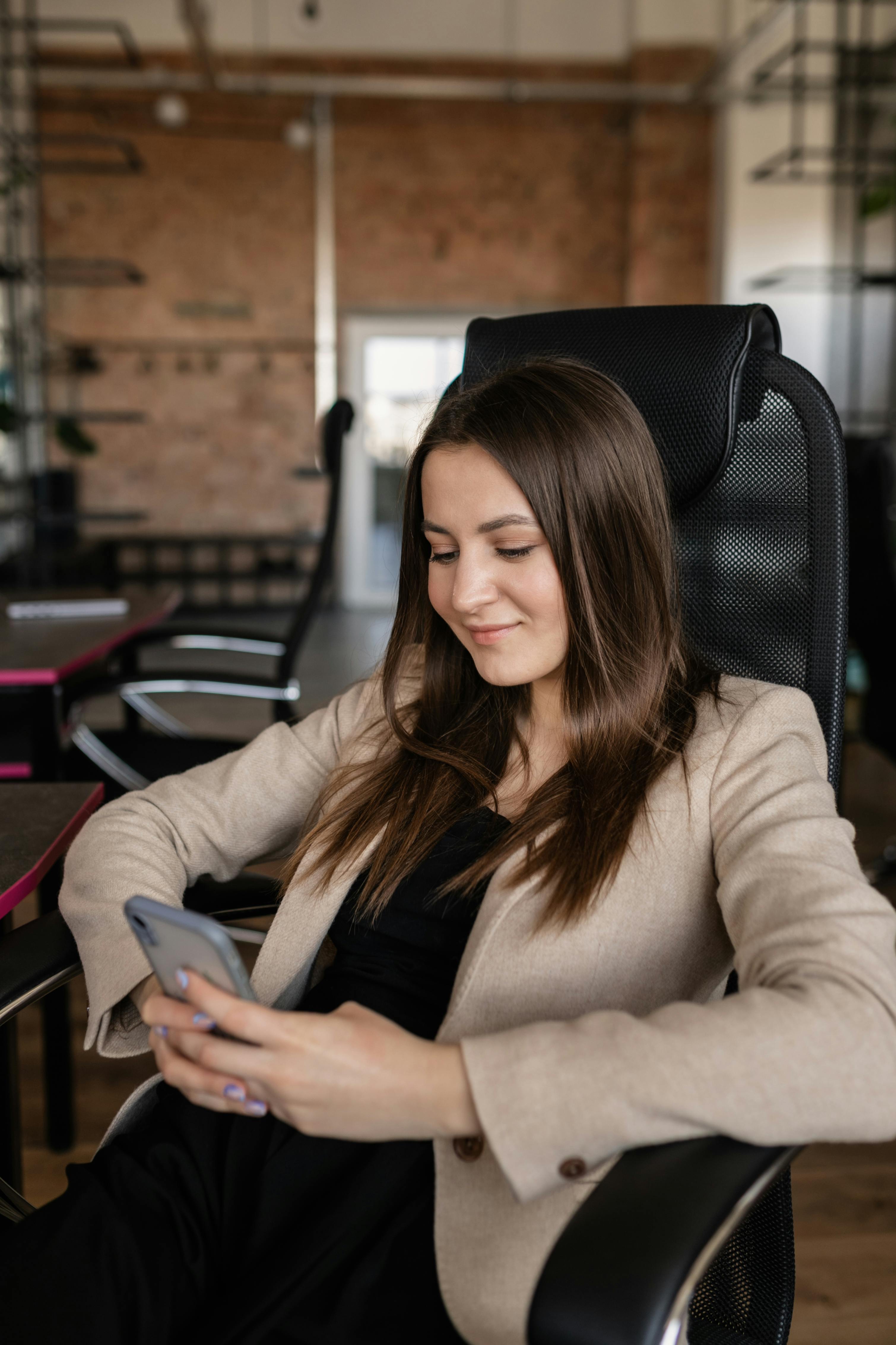 a woman in beige blazer sitting on the chair while using her mobile phone