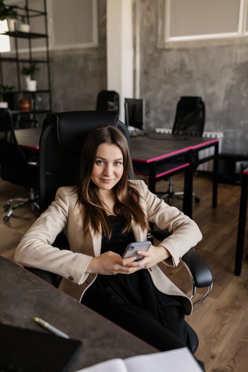Free Confident woman sitting with phone in modern open office, relaxed atmosphere. Stock Photo