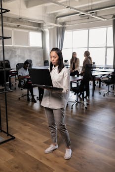 Asian woman standing in a modern office, focusing on her laptop amidst coworkers.