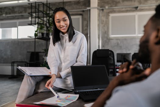 Asian woman in white long sleeves engages with a colleague in a modern workspace. Office collaboration.