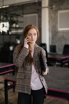 Professional woman making a phone call in a modern office environment, holding a laptop.