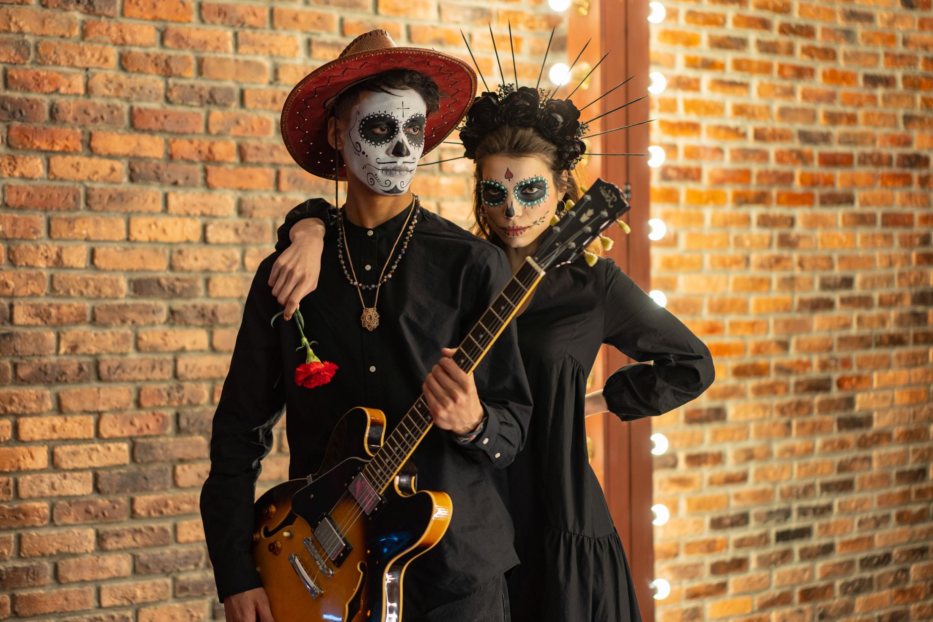Couple celebrating Day of the Dead with face paint and guitar in front of a brick wall.