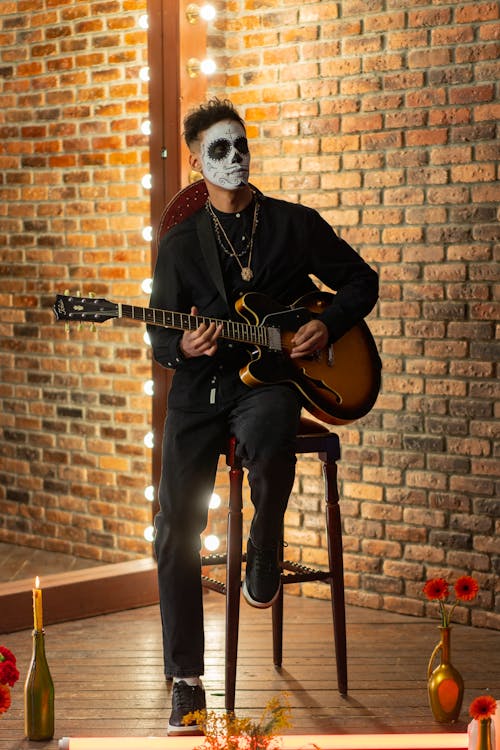 A Man with Face Paint Sitting on a Chair while Playing a Guitar