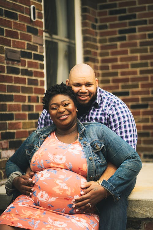 Cheerful black man touching tummy of pregnant African American female while looking at camera together near brick building on terrace