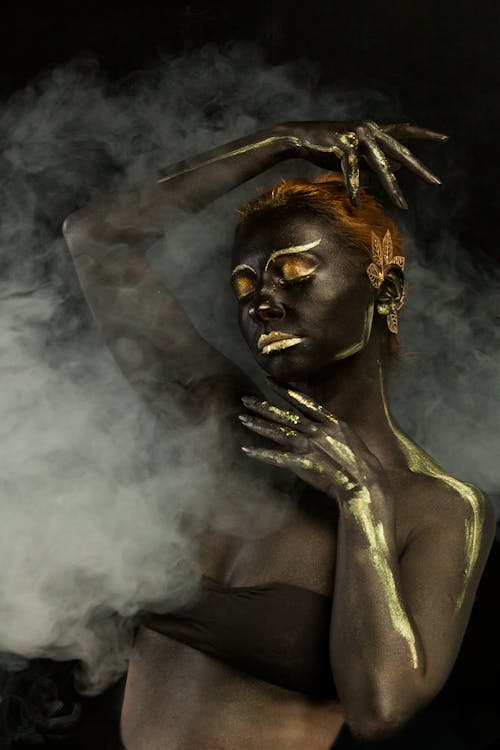 Free Young female in bra with body art covered with dark paint and golden pigment over face and hands standing against black background in white smoke with closed eyes and raised arm Stock Photo