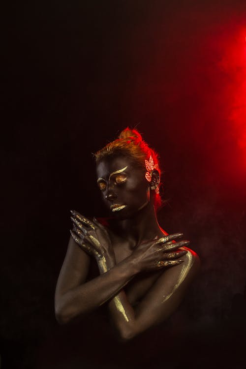 Young sensual female with black and golden body art and closed eyes covering breast while standing against black background in red neon illumination