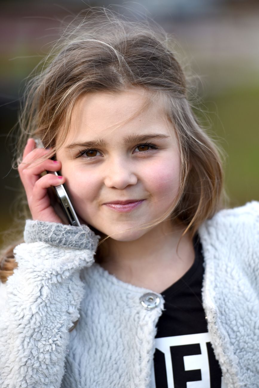 A young girl smiles while talking on a mobile phone outdoors in natural light.