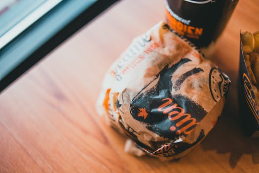 From above of delicious burger wrapped in craft paper served on wooden table with plastic cup of soda and fries