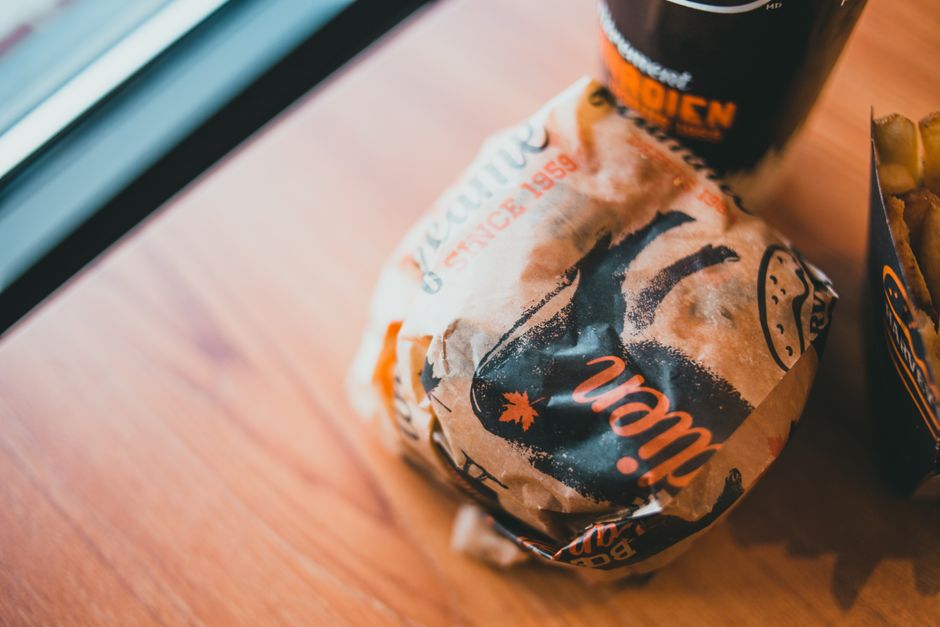 From above of delicious burger wrapped in craft paper served on wooden table with plastic cup of soda and fries