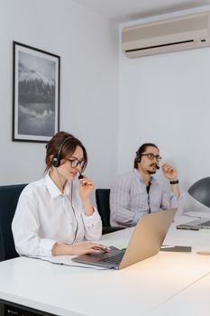 Two professionals working remotely in a modern office space, wearing headsets and using laptops.