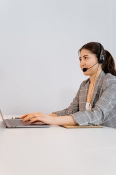 Professional woman in plaid blazer using laptop with headset in an office setting.