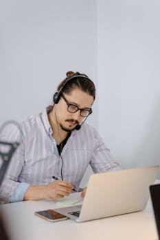 Focused man with headset taking notes and working on a laptop in an office setting.